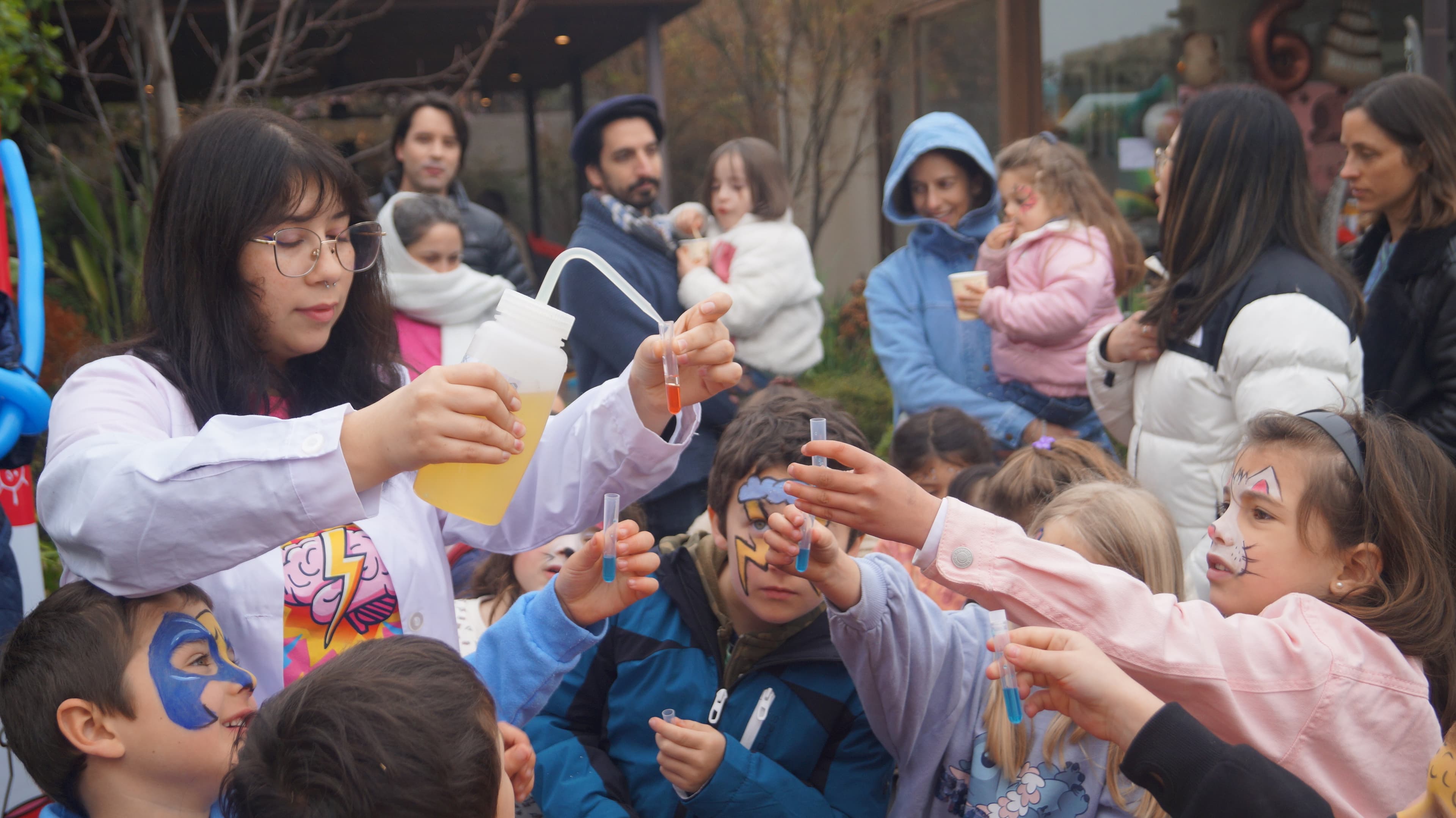 Educadora con bata vierte un líquido en un tubo de ensayo mientras niños con la cara pintada extienden sus tubos