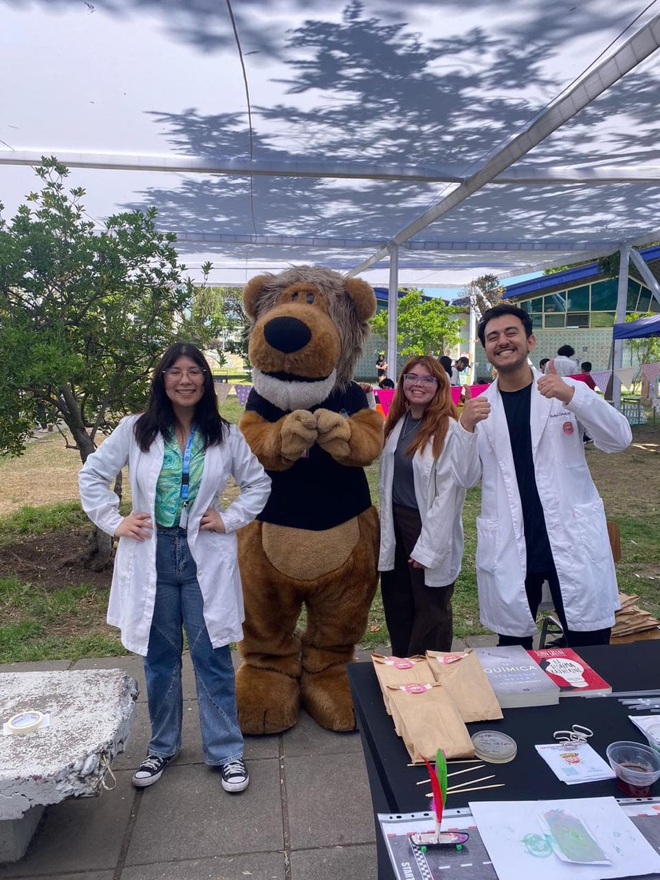 Tres científicos con bata blanca posan junto a una mascota de peluche en un evento al aire libre